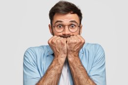 Nervous frightened male keeps fists near mouth, has worried expression, stares at camera, isolated over white background. Axious unshaven young man feels depressed. People and concern concept