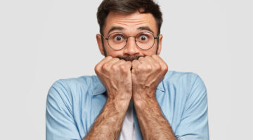 Nervous frightened male keeps fists near mouth, has worried expression, stares at camera, isolated over white background. Axious unshaven young man feels depressed. People and concern concept
